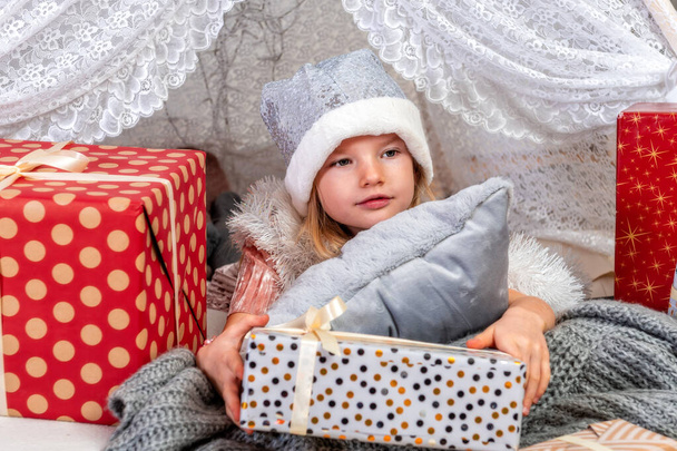 portrait of a cute little girl child wearing a silver color Christmas hat lying between gift boxes, Christmas concept - Photo, image