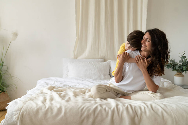 Poika syleilee hymyilevää äitiä aamulla, istuu sängyllä yöpuvussa kotona viikonloppuna. Perherakkaus - Valokuva, kuva