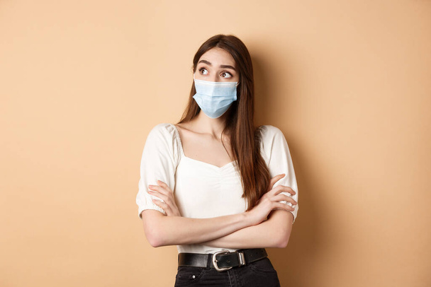 Koncepcja pandemii i opieki zdrowotnej. Przemyślana dziewczyna w masce na twarzy patrząc na puste logo przestrzeni, stojąc ciekawa na beżowym tle - Zdjęcie, obraz