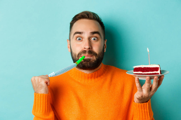 Close-up of funny adult man celebrating his birthday, holding bday cake with candle, blowing party wistle and rejoicing, standing over light blue background - Foto, Bild