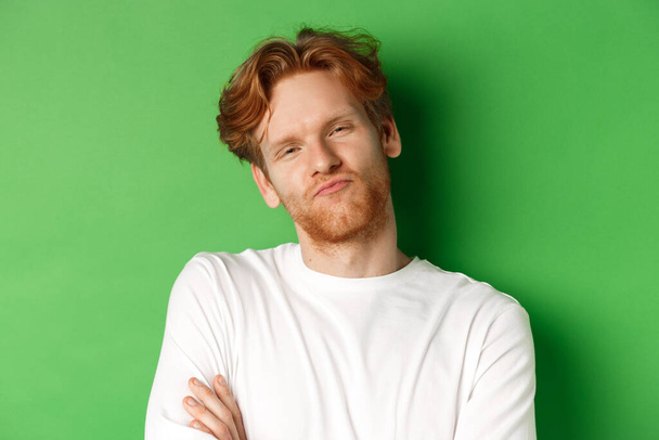 Emotions and fashion concept. Close-up of skeptical young man with red hair and beard pucker lips, looking at camera as waiting for something, standing over green background - Photo, Image
