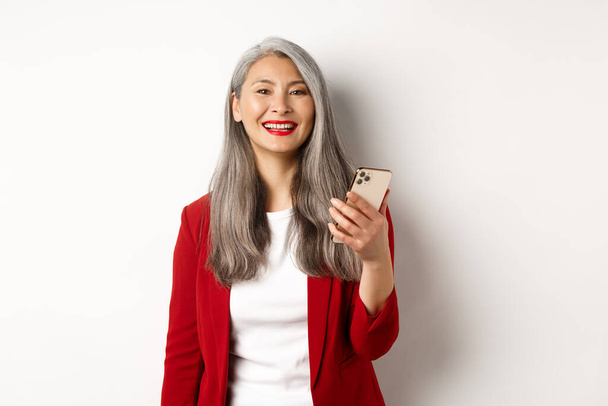 Asian female entrepreneur in red blazer using smartphone, smiling happy at camera, standing over white background - Photo, Image