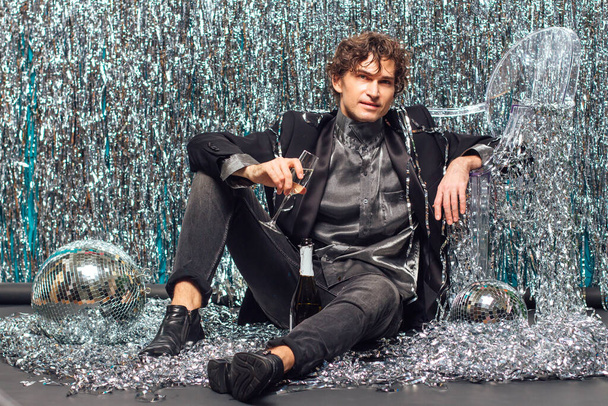 Tall attractive man sitting on the floor over the sparkle background and mirror disco ball holding glass of champagne. Party concept. - Fotó, kép