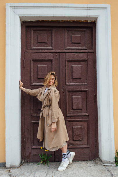 Young smiling millennial woman with wild hair dressed in an autumn coat posing near the door of an old building. - Zdjęcie, obraz
