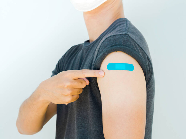 Vaccinations, bandage plaster on vaccinated people's arm concept. Blue color adhesive bandage on man's arm who wearing white face mask, pointing own shoulder after vaccination treatment. - Fotó, kép