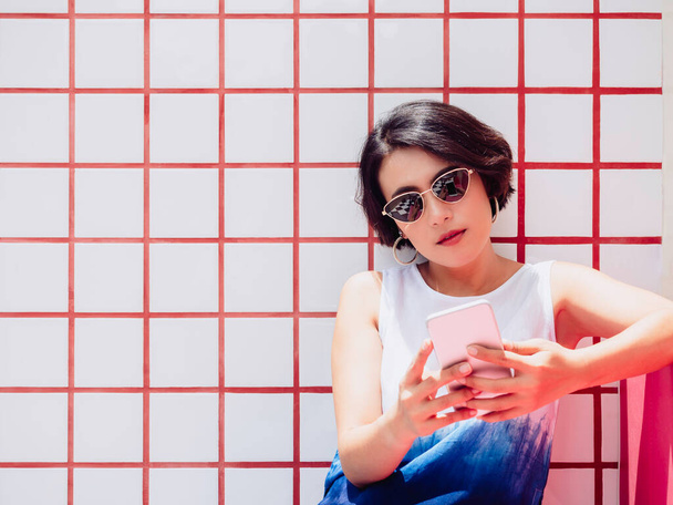 Ελκυστική κομψή ασιατική γυναίκα κοντά μαλλιά φορώντας λευκό πουκάμισο και γυαλιά ηλίου κάθεται σε κόκκινο κάθισμα και χρησιμοποιώντας smartphone σε λευκό και κόκκινο πλακάκια φόντο στον τοίχο με αντίγραφο χώρο. - Φωτογραφία, εικόνα