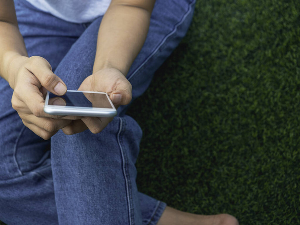 Close-up mobile phone in hands of woman wearing white sleeveless shirt and blue jeans sitting with relax on green artificial grass background with copy space. Hands holding a smartphone. - Valokuva, kuva