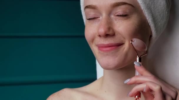 Urocza kobieta masuje swoją piegowatą twarz wałkiem do skóry uśmiechniętym w domu - Materiał filmowy, wideo