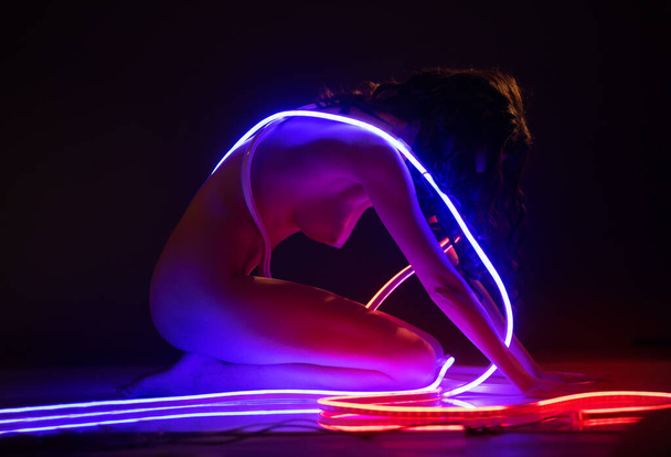 Een slank, mooi, naakt meisje poseert op een donkere achtergrond met een neon lint in haar handen. - Foto, afbeelding