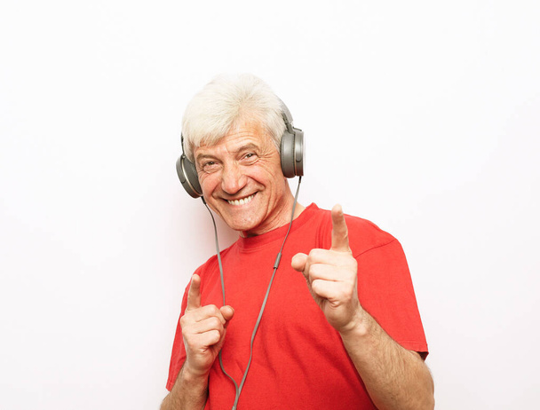 Lifestyle, tehnology and old people concept: Handsome old man in casual wear and headphones is listening to music and show ok sign on white background - Fotó, kép