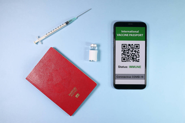 Digitaalinen kansainvälinen rokotepassi älypuhelin näyttö qr-koodi app coronavirus covid 19 immunisaatio punainen passi kirja pieni pullo kertakäyttöinen lääketieteellinen ruisku neula valo taivaansininen tausta - Valokuva, kuva