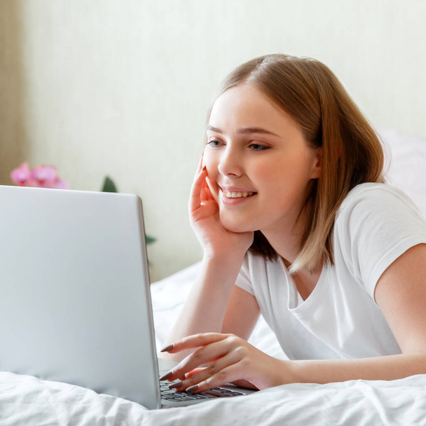 Młoda kobieta uśmiecha się i pracuje na laptopie leżąc rano w łóżku w domu. Szczęśliwa dziewczyna w piżamie portret studiuje zakupy online lub planowania jej dzień korzystania z laptopa w sypialni wnętrza. - Zdjęcie, obraz