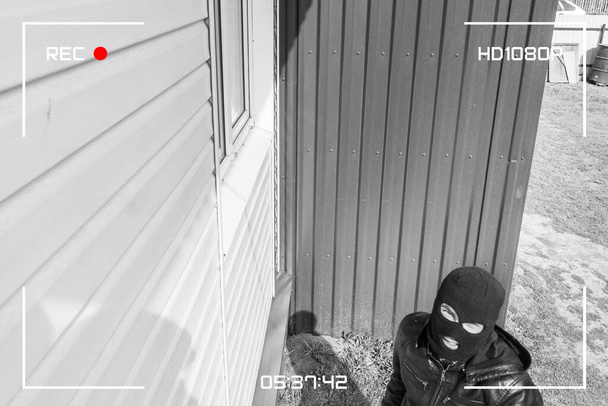 A man in a black mask is trying to rob a house. There is a recording on an outdoor video surveillance camera. The robber noticed a security camera. Robbery of a private house. Criminal concept - Zdjęcie, obraz