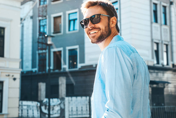 Portrait of handsome smiling stylish hipster lambersexual model.Modern man dressed in blue shirt. Fashion male posing near skyscraper on the street background in sunglasses. Outdoors at sunset  - Zdjęcie, obraz