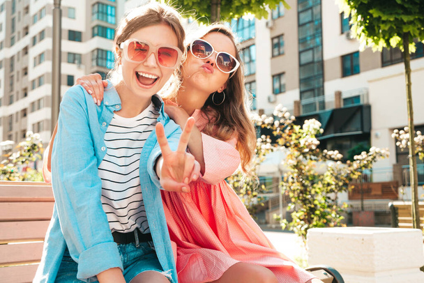 Zwei junge schöne lächelnde Hipsterinnen in trendigen Sommerkleidern und Kleidern. Sexy unbeschwerte Frauen posieren auf der Straße. Positiv reine Models, die bei Sonnenuntergang auf einer Bank sitzen und Spaß haben - Foto, Bild