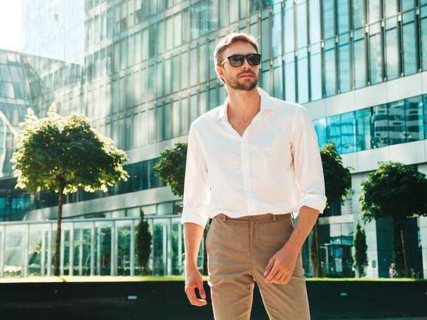 Portrait of handsome smiling stylish hipster lambersexual model.Modern man dressed in white shirt. Fashion male posing in the street background near skyscrapers in sunglasses - Fotoğraf, Görsel