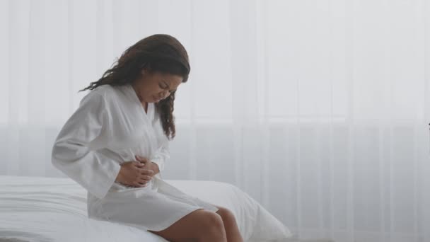 Douleurs menstruelles. Jeune femme afro-américaine souffrant de douleurs abdominales aiguës, assise sur le lit, espace vide - Séquence, vidéo