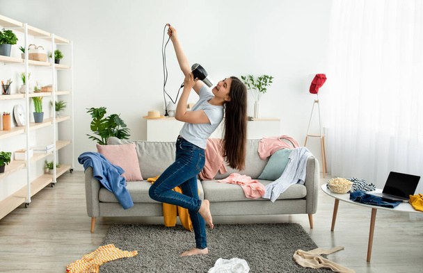 Indian teenage girl dancing and singing in messy room, using blowdryer as microphone, giving domestic concert - Foto, Bild