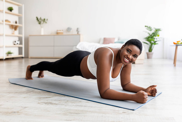 Urheilu ja laihtuminen käsite. Ylipainoinen afroamerikkalainen nainen tekee harjoituksia matolla kotona, täyspitkä - Valokuva, kuva