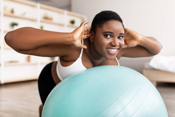 Χαρούμενη συν μέγεθος μαύρη γυναίκα κάνει ασκήσεις με μπάλα γυμναστικής, ενίσχυση των μυών της πλάτης στο σπίτι - Φωτογραφία, εικόνα