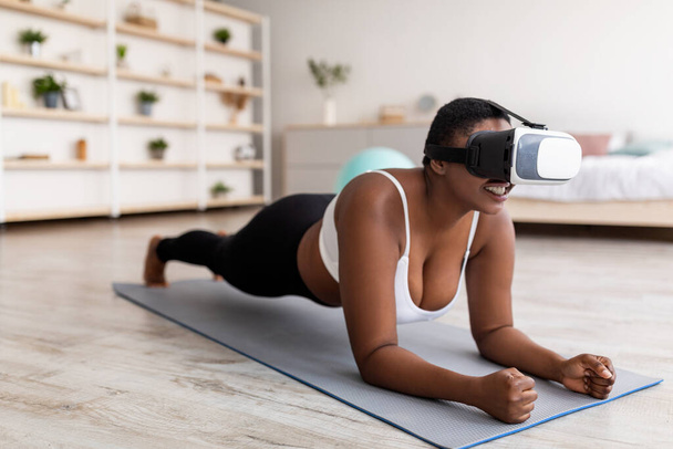 Domestic virtual training. Curvy young black lady in VR glasses standing elbow plank at home - 写真・画像