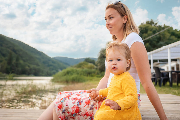 夏だ。桟橋に座っている母親と赤ん坊の女の子。サイドビュー。家族の休暇の概念. - 写真・画像