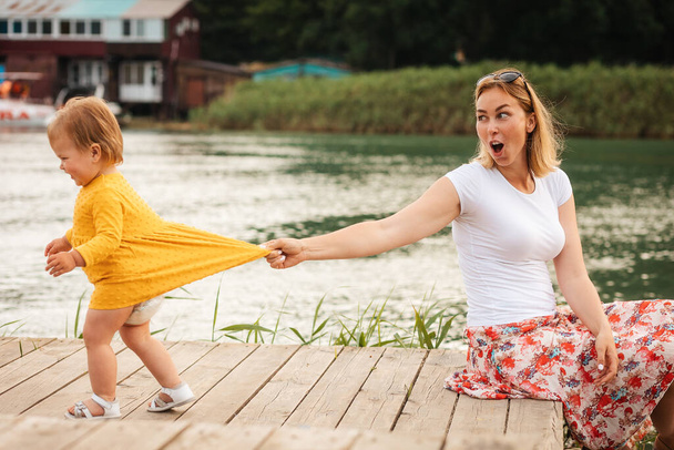 Sommerzeit. Junge kaukasische Mutter und ihre kleine Tochter spielen auf der Seebrücke am Fluss. Seitenansicht. Das Konzept der Kindererziehung. - Foto, Bild