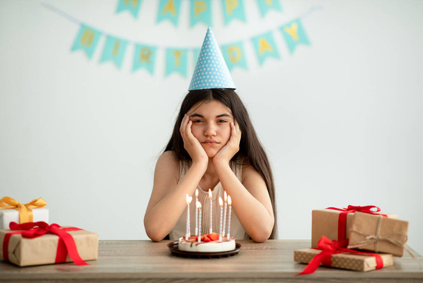 Muotokuva pettynyt Intian teini tyttö puolue hattu istuu yksin pöydässä lahjoja ja syntymäpäivä kakku - Valokuva, kuva