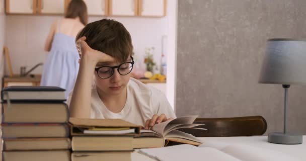 Junge liest Bücher, während seine Mutter in der Küche kocht. Kind macht Hausaufgaben. - Filmmaterial, Video