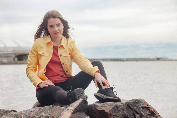 Pretty young sexy woman of athletic figure in bright clothes walking on coastline in city. Walk in city sea beach. Portrait of cute pretty female in yellow jacket and black pants on sea coast - Φωτογραφία, εικόνα