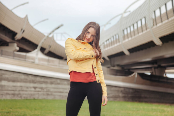 Pretty young sexy woman in clothes standing against background of road junction. Walking in city street. Portrait of cute pretty female in yellow jacket and black pants on street way - Zdjęcie, obraz