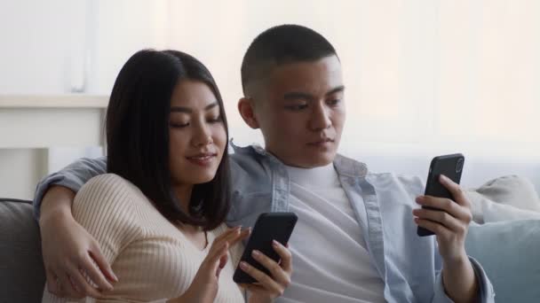 Κινεζικό ζευγάρι που χρησιμοποιεί κινητά τηλέφωνα κάθεται στον καναπέ στο σπίτι - Πλάνα, βίντεο