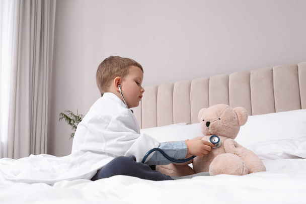 Cute little boy in pediatrician's uniform playing with stethoscope and toy bear at home - 写真・画像