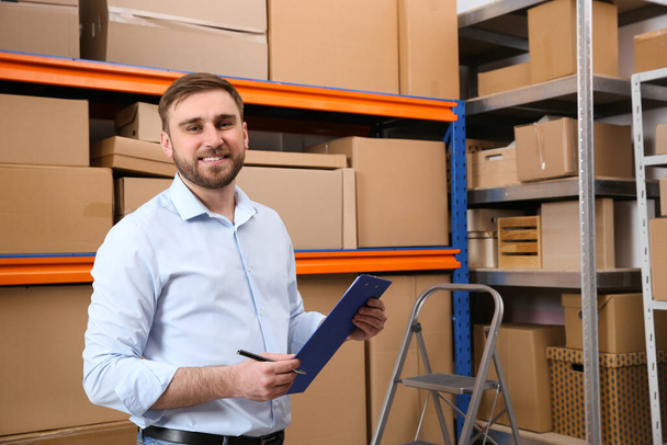Young businessman with clipboard near rack of cardboard boxes at warehouse - Foto, Imagen