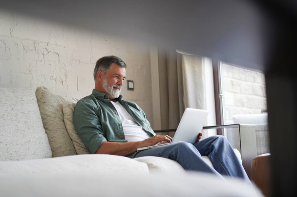 Portrait of happy mature man in casual clothes using laptop lying on sofa in house. - Φωτογραφία, εικόνα