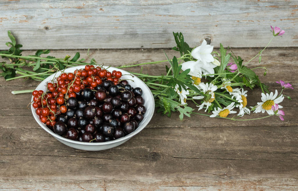 A plate with black and red currants next to a delicate bouquet of meadow flowers: chamomiles and bells on the wooden steps of an old house. Summer holidays concept. Still life. Summer time - Foto, Bild