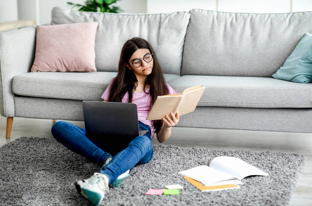 Indien adolescent fille lecture manuel, assis sur le sol avec ordinateur portable et matériel d'étude, apprendre à distance de la maison - Photo, image