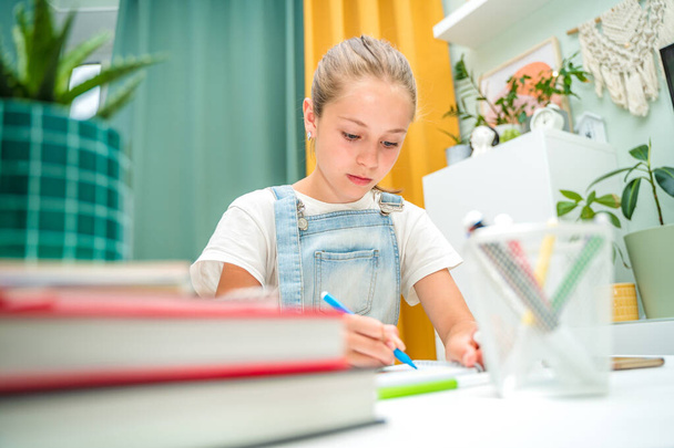 Ένα μικρό κορίτσι ζωγραφίζει με στυλό τσόχας στο σπίτι σε ένα μοντέρνο σαλόνι. Ολοκλήρωση της εργασίας με τη βοήθεια των βιβλίων - Φωτογραφία, εικόνα