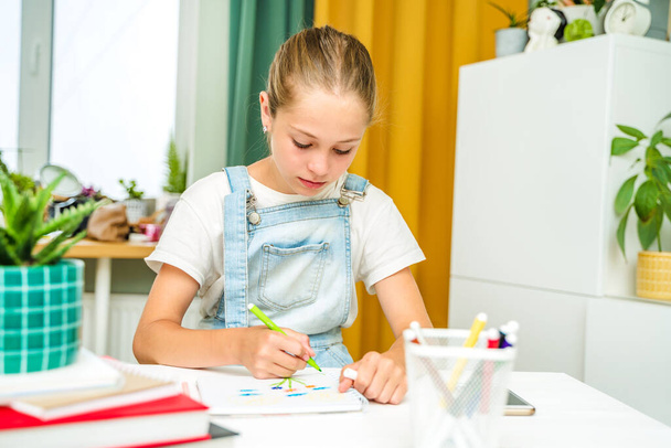 Ein kleines Mädchen zeichnet mit Filzstiften zu Hause in einem modernen Wohnzimmer. Hausaufgaben mit Hilfe von Büchern erledigen - Foto, Bild