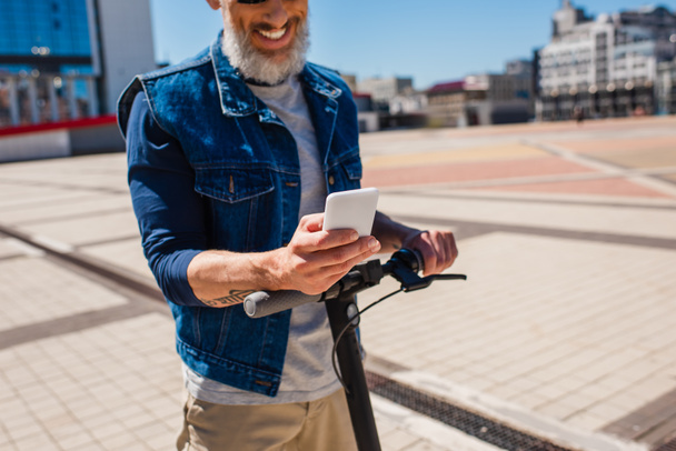 обрезанный вид счастливого и зрелого мужчины с помощью смартфона во время езды на электронном скутере снаружи  - Фото, изображение