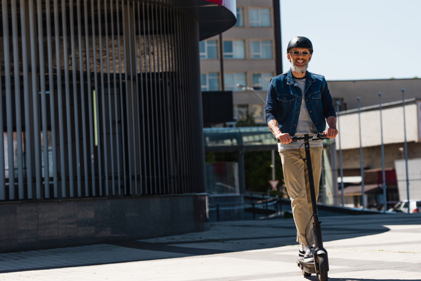 full length of pleased man in sunglasses and helmet riding e-scooter in urban city  - Φωτογραφία, εικόνα