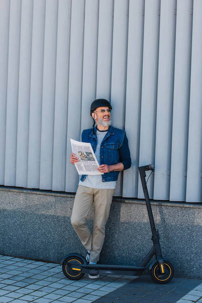 full length of cheerful and mature hipster in helmet and sunglasses holding newspaper near e-scooter  - Photo, Image