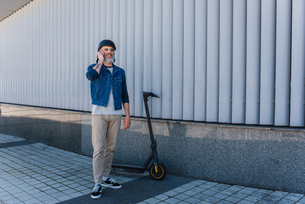 full length of cheerful and mature hipster in helmet and sunglasses talking on smartphone near e-scooter  - 写真・画像