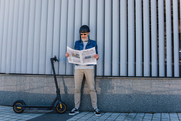 Reifer Hipster mit Helm und Sonnenbrille liest Reisezeitung in der Nähe von E-Scooter  - Foto, Bild