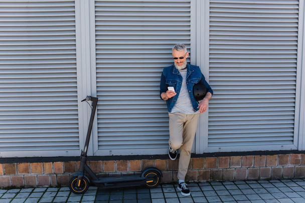 full length of happy and mature man in sunglasses using smartphone near e-scooter  - Valokuva, kuva