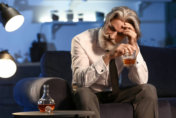 пьяный пожилой мужчина со стаканом виски поздно вечером дома - Фото, изображение