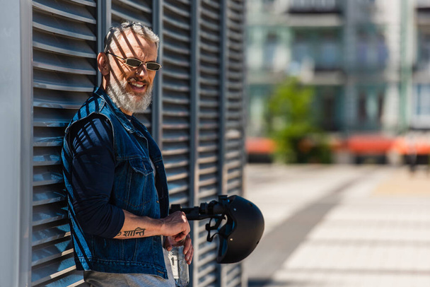 bearded middle aged man in sunglasses smiling and holding bottle of water near e-scooter with helmet - Φωτογραφία, εικόνα