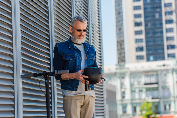 middle aged man in sunglasses smiling and holding helmet near electric scooter  - Foto, Imagem