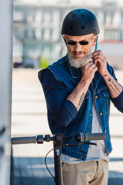 tattooed middle aged man in sunglasses wearing helmet near electric scooter  - 写真・画像
