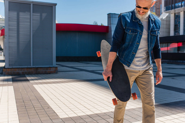 bearded middle aged man in sunglasses holding longboard on urban street - 写真・画像